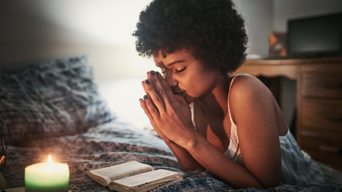 Mujer orando sobre la cama con una biblia y una vela.